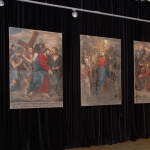 Trzy obrazy przedstawiające sceny Drogi Krzyżowej wiszą na czarnej ścianie